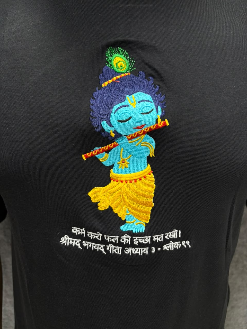 Close up of embroidered bal Krishna with flute and peacock feather on black cotton t-shirt with bagwadgita slok