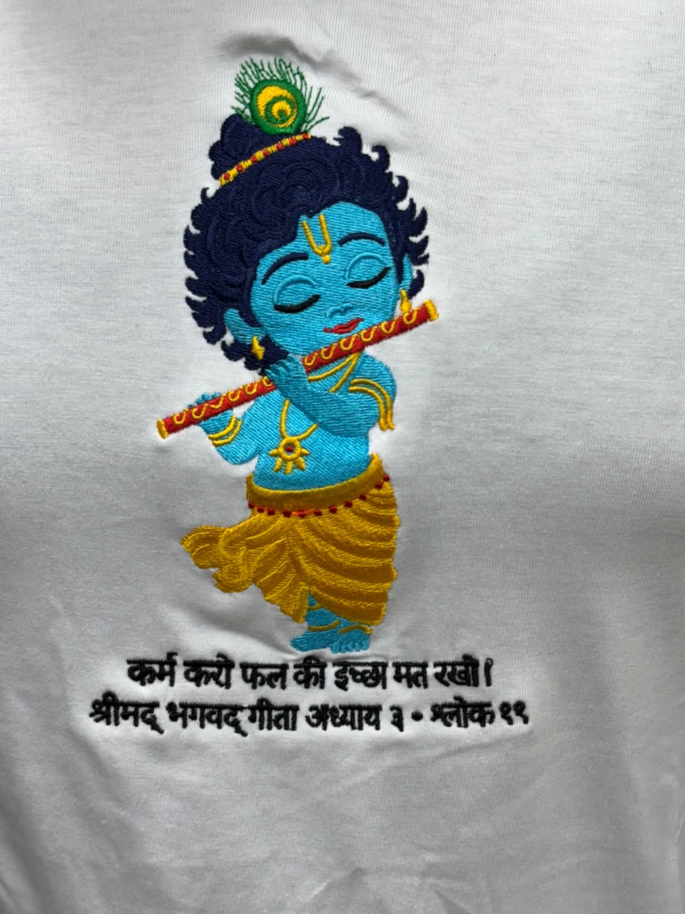 Close up of embroidered bal Krishna with flute and peacock feather on white cotton t-shirt with bagwadgita slok