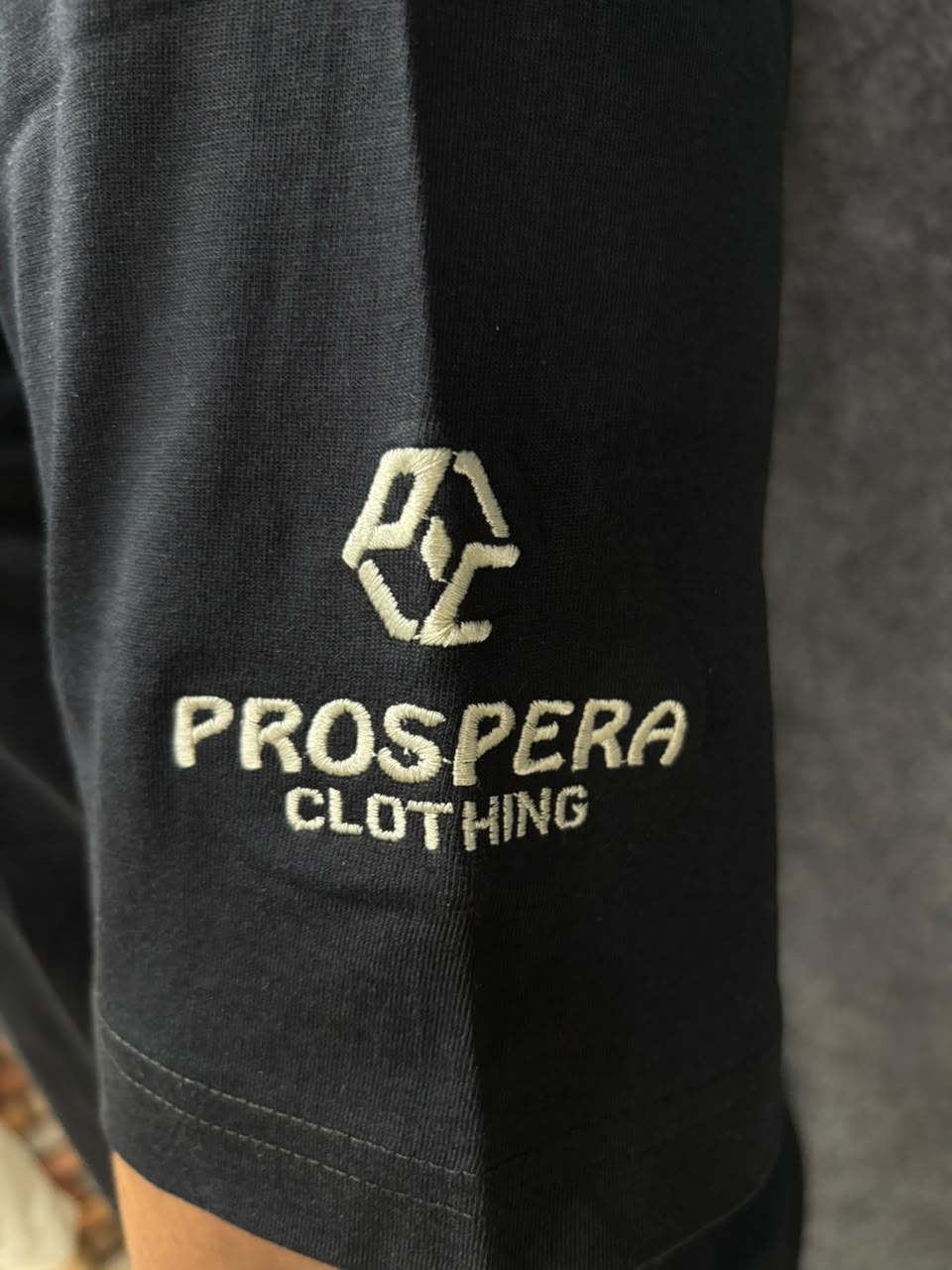 Close-up of a black garment sleeve with a gold embroidered logo and the text PROSPERA CLOTHING